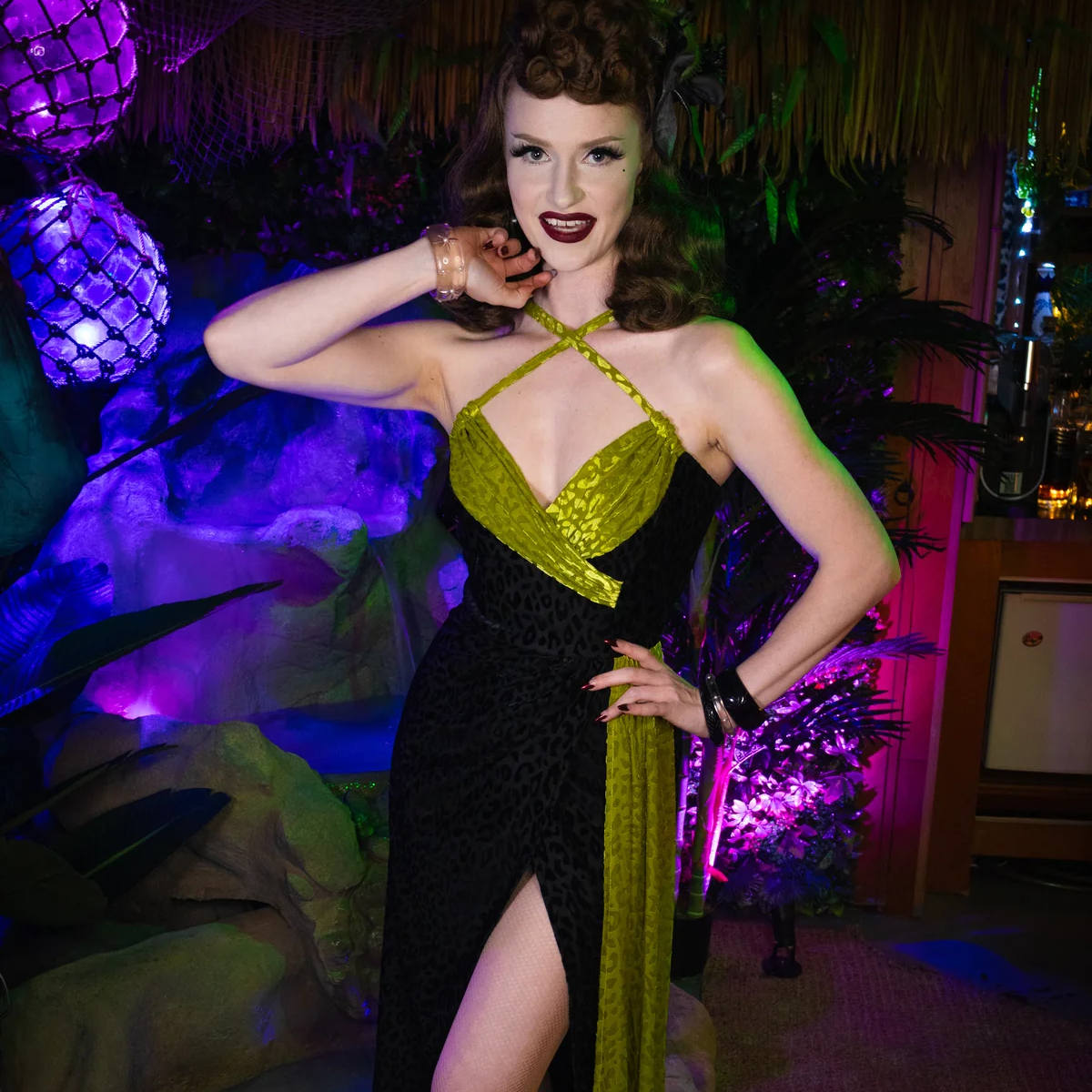 A woman posing in a retro 1950s-style halter dress with a black leopard print skirt and a shiny olive green bodice and side panel, set against a tropical-themed party background with purple lighting.