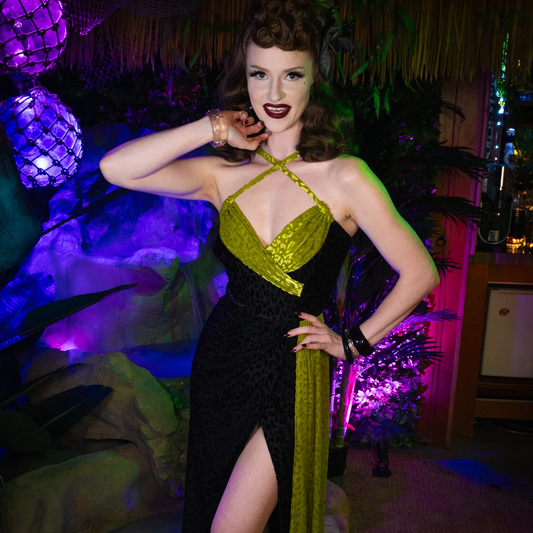 A woman posing in a retro 1950s-style halter dress with a black leopard print skirt and a shiny olive green bodice and side panel, set against a tropical-themed party background with purple lighting.