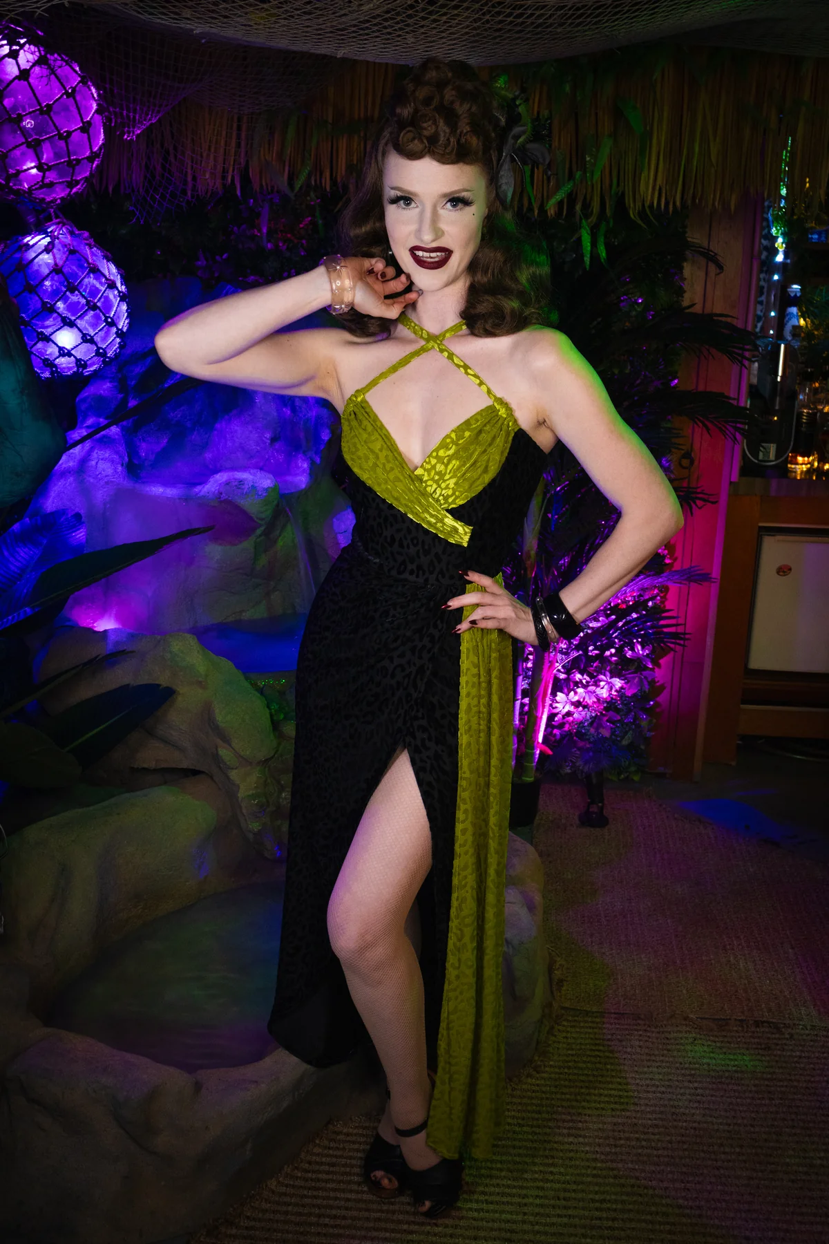 A woman posing in a retro-style halter dress with a black leopard print skirt and a shiny olive green bodice, set against a tropical-themed dimly lit background with purple lighting.