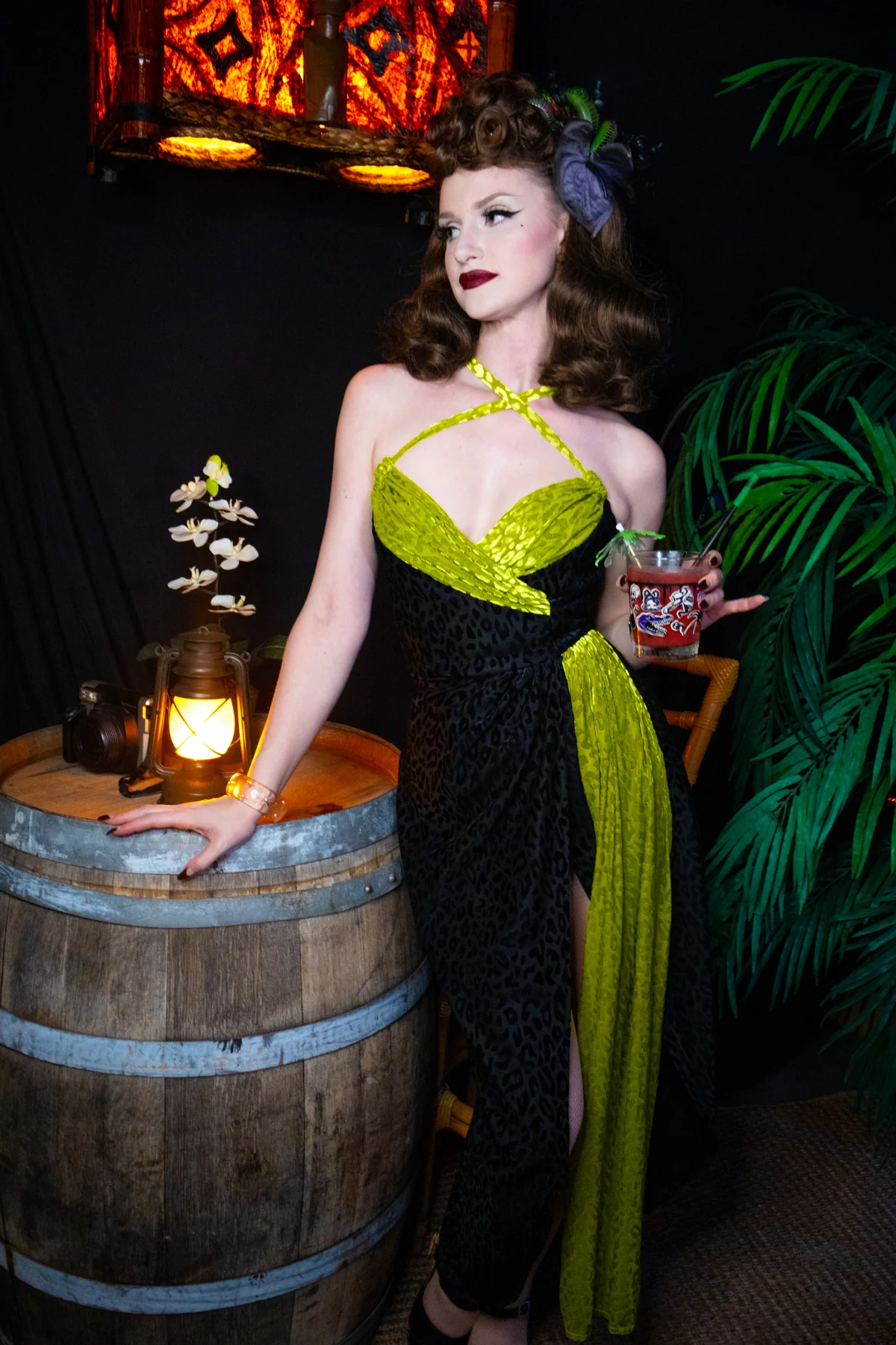 A woman models a 1950s-style halter dress with a black leopard print and bright lime green sequined panels, posing next to a wooden barrel in a dimly lit, retro-themed setting.