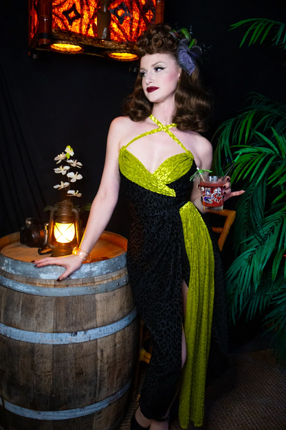 A woman models a 1950s-style halter dress with a black leopard print and bright lime green sequined panels, posing next to a wooden barrel in a dimly lit, retro-themed setting.