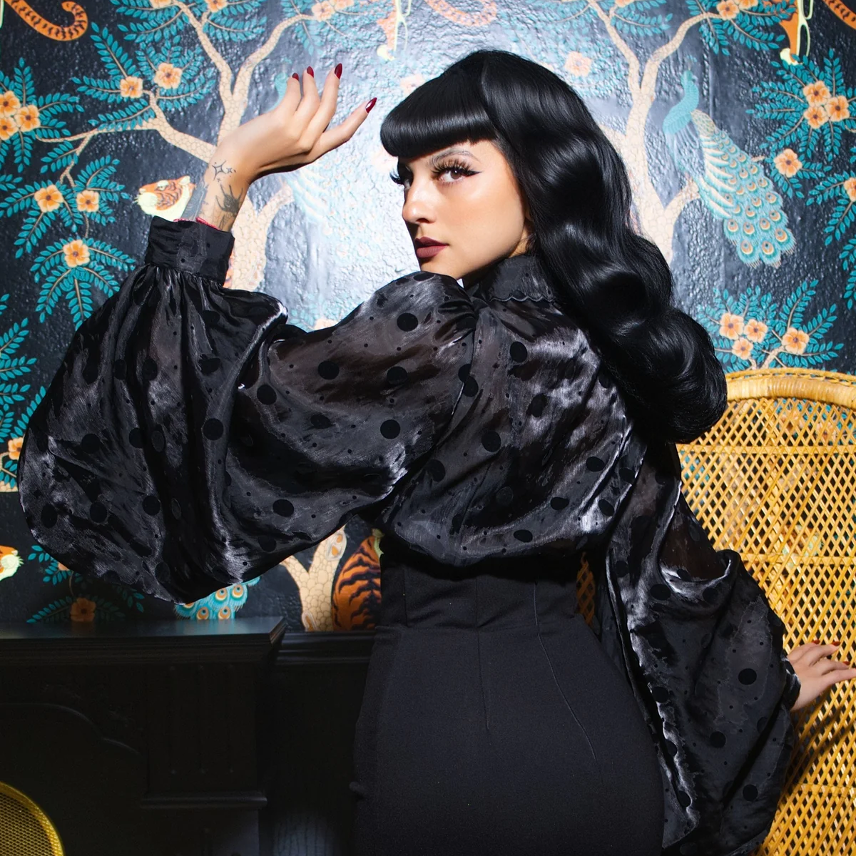 A woman posing in a black velvet and satin two-piece outfit with dramatic flared sleeves and a flared skirt, standing in front of a decorative tiger-print wallpaper background next to a yellow wicker chair.