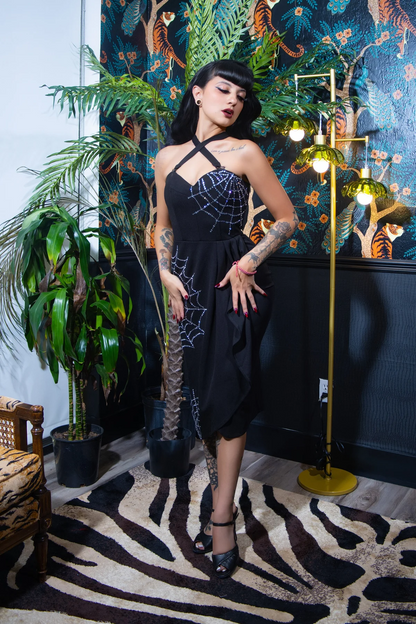 A woman models a black spaghetti-strap dress with white spiderweb patterns on the bodice and skirt, standing in front of a dark patterned wallpaper and a gold peacock mirror.