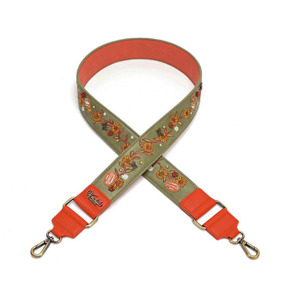 A fabric shoulder strap with an olive green background featuring a colorful embroidered floral and pumpkin pattern, finished with bright orange-red leather ends and gold-tone hardware.