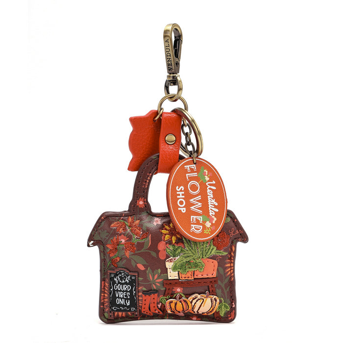 A decorative autumn-themed leather keychain shaped like a house, featuring a floral and pumpkin pattern in earthy tones. It has a bright orange-red bow, a brass clasp, and two hanging tags: one oval-shaped tag reading 'Flower Shop' and one small black sign that says 'GROUNDS VIBES ONLY'.