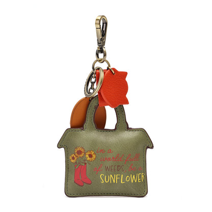 An olive green leather keychain shaped like a house, featuring a printed design with sunflowers, red boots, and the phrase 'in a world full of weeds, I'm a sunflower' in yellow and pink. It includes a gold-tone lobster clasp, key ring, and orange and brown leather accents.