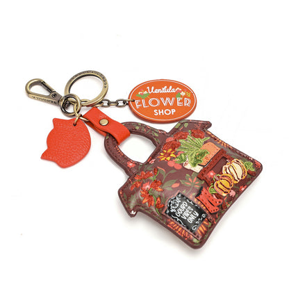 A decorative tote bag keychain with a floral print, attached to a metal clasp and key ring. It includes an orange leather flower-shaped charm and an oval orange tag reading 'Montita FLOWER SHOP'.