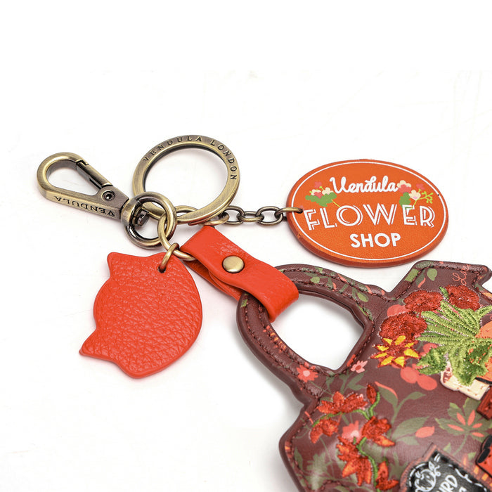 A close-up of a handbag keychain featuring a terracotta orange textured leather flower-shaped charm, a matching orange oval key tag with 'Ventuela Flower Shop' text, and a brown floral-patterned handbag strap.