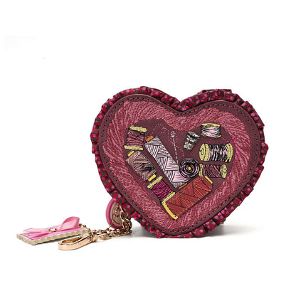 A heart-shaped coin purse with a pink and burgundy ruffled border, featuring a central patchwork design of sewing thread spools and fabric squares. It includes a small pink bow charm and a gold-tone keychain ring on a white background.