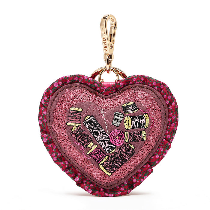 A heart-shaped embroidered pouch with a floral ruffled border, featuring a central patch of pink and purple embroidery with thread spools and a gold-tone carabiner clip.