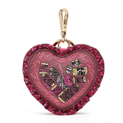 A heart-shaped embroidered pouch with a floral ruffled border, featuring a central patch of pink and purple embroidery with thread spools and a gold-tone carabiner clip.