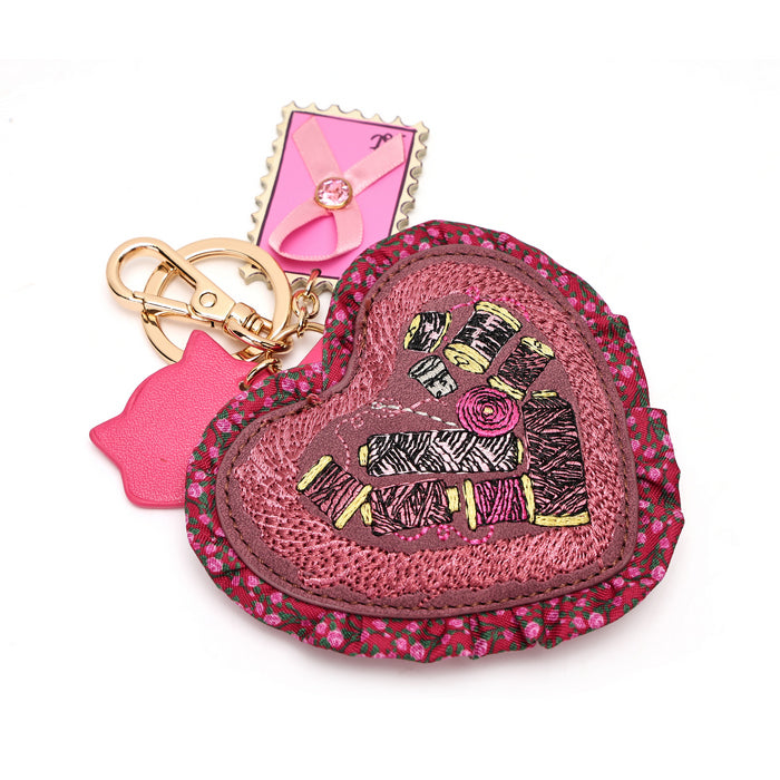 A heart-shaped pink leather keychain bag with a floral border, a central patch of black and pink thread embroidery, a pink flower applique, a gold-tone key ring, and a pink cat-shaped charm, shown on a white background.