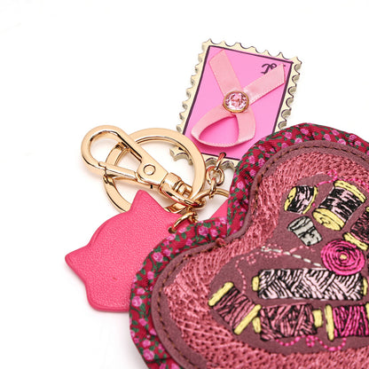 A pink heart-shaped embroidered keychain bag with a gold-tone clasp, a pink cat-shaped charm, and a decorative pink tag on a white background.
