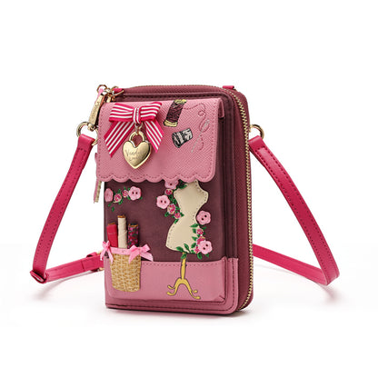 A pink and burgundy crossbody wallet bag with decorative fashion-themed appliques, including a ribbon bow, a gold heart charm, a basket pocket, and a mannequin silhouette, shown on a white background.