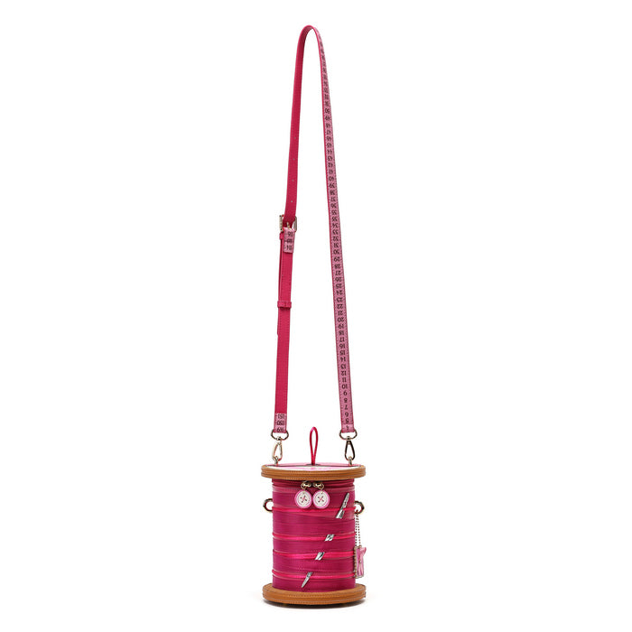 A bright pink spool-shaped crossbody bag with a matching pink strap printed with a white measuring tape design, set against a plain white background.