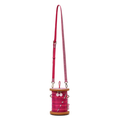 A bright pink spool-shaped crossbody bag with a matching pink strap printed with a white measuring tape design, set against a plain white background.