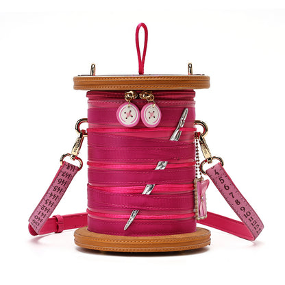 A bright pink spool-shaped crossbody bag with a matching pink strap printed with a white measuring tape design, set against a plain white background.