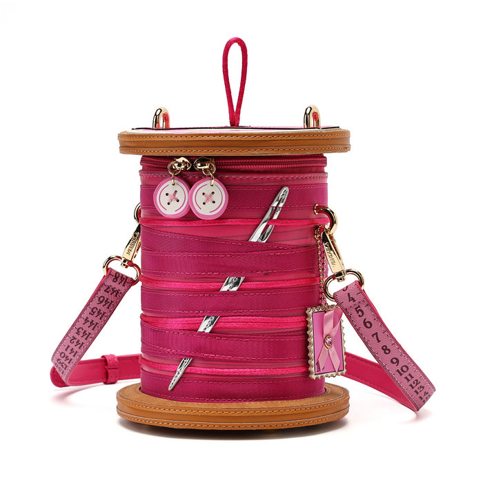 A pink spool-shaped shoulder bag with a tan faux-leather rim, pink zipper details, decorative buttons, and a pink strap printed with text, shown on a white background.