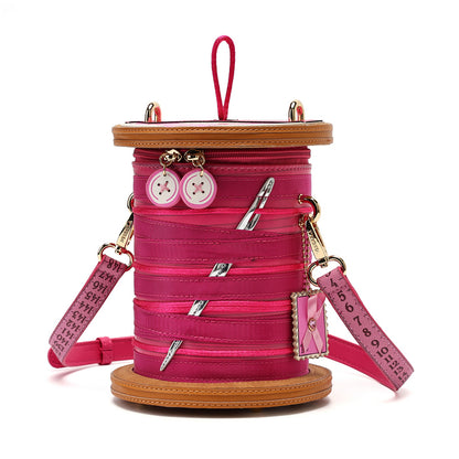 A pink spool-shaped shoulder bag with a tan faux-leather rim, pink zipper details, decorative buttons, and a pink strap printed with text, shown on a white background.