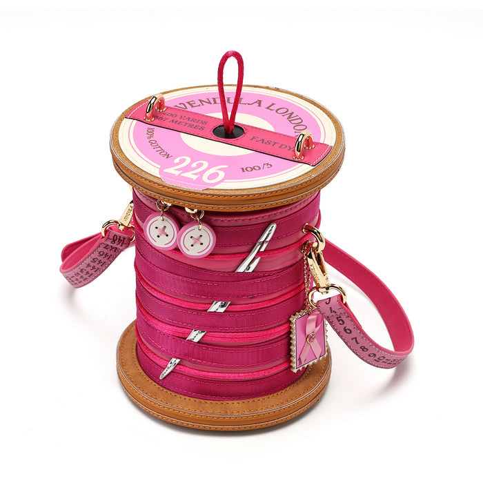 A pink spool-shaped shoulder bag designed to look like a sewing thread spool, featuring a pink strap, decorative buttons, and a hanging charm, photographed on a white background.