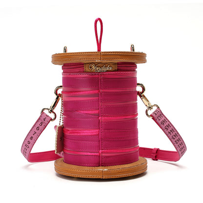 A pink faux leather drum-shaped crossbody bag with tan leather trim, a top carrying loop, and a pink strap printed with black measurement markings, shown on a white background.