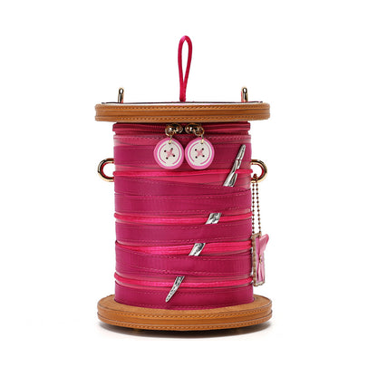 A novelty spool-shaped handbag in bright pink faux leather with tan leather trim, featuring decorative pink buttons, silver nails, and a pink zipper pull on a white background.