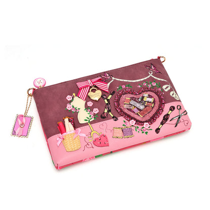 A pink and mauve zippered clutch bag decorated with various sewing-themed appliques, including a heart-shaped patch, scissors, a quill pen, and a small charm, shown on a plain white background.