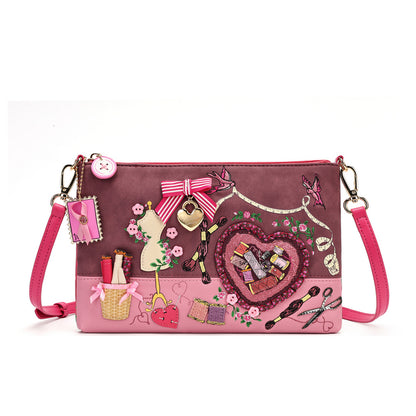 A pink and mauve crossbody clutch bag decorated with various sewing-themed charms, including a heart-shaped patch, ribbon bows, scissors, and a dress form, shown on a plain white background.