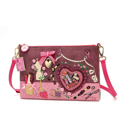 A pink and mauve crossbody clutch bag decorated with various sewing and craft-themed charms, including a ribbon bow, scissors, a heart patch, and a miniature dress form, on a plain white background.