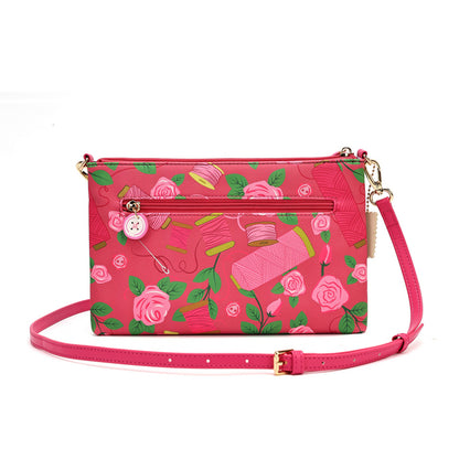 A pink crossbody handbag with a floral and sewing-themed print featuring roses, thread spools, scissors, and buttons. The bag has a pink leather strap, gold-tone hardware, and a front zippered pocket.