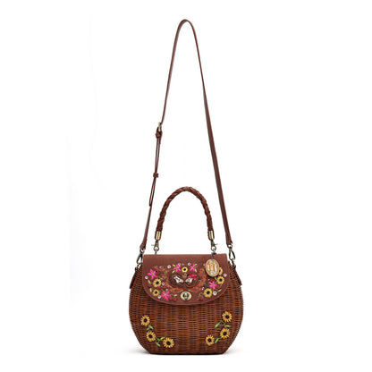 A brown woven wicker shoulder bag with a long shoulder strap, top handle, and floral embroidered details on the front flap and body, shown against a plain white background.