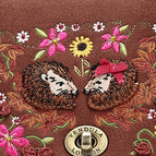 A close-up of a brown embroidered textile featuring two hedgehogs surrounded by pink flowers and a red sunflower, with a circular gold-toned emblem near the bottom center.