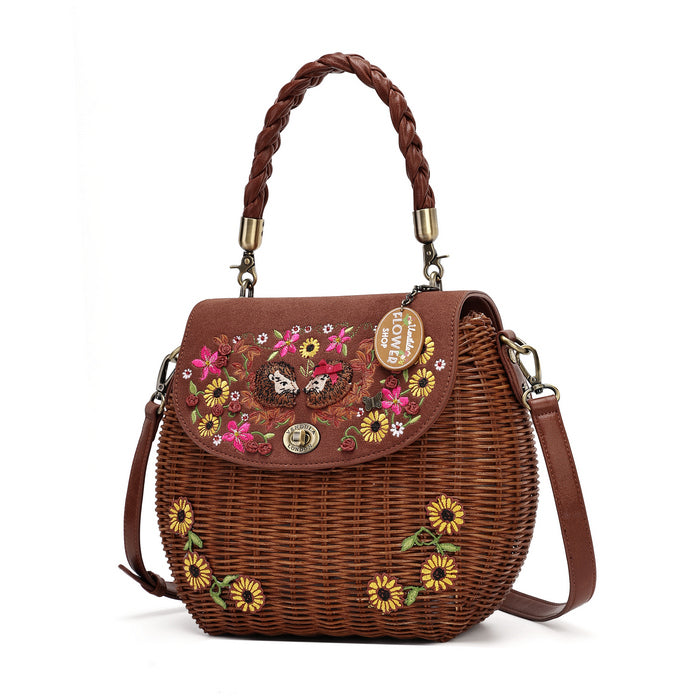 A brown woven wicker handbag with a braided top handle, a detachable shoulder strap, and colorful floral embroidery on the front flap. The bag features a gold-tone turn-lock closure and a hanging charm that reads 'LOVE IS LONELY'.