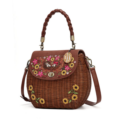 A brown woven wicker handbag with a braided top handle, a detachable shoulder strap, and colorful floral embroidery on the front flap. The bag features a gold-tone turn-lock closure and a hanging charm that reads 'LOVE IS LONELY'.
