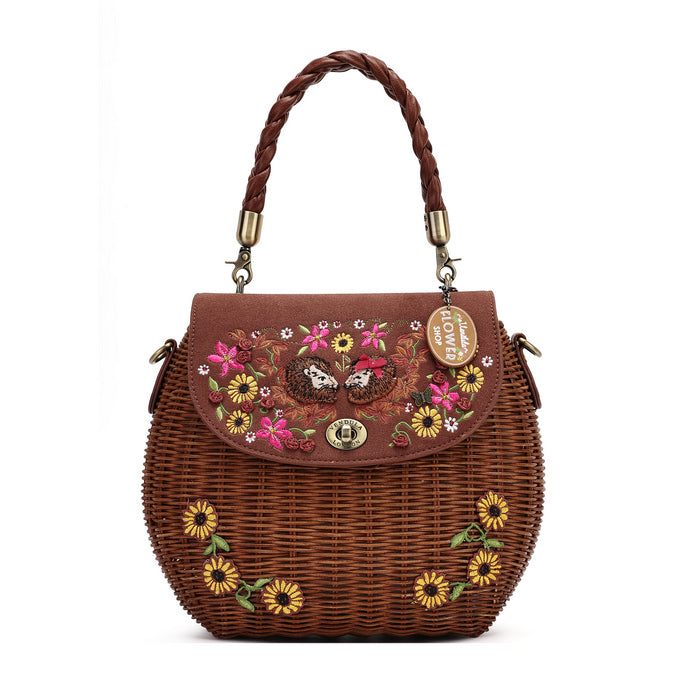A brown woven wicker handbag with a braided top handle, featuring a floral embroidered flap and yellow sunflower details on the front, set against a plain white background.