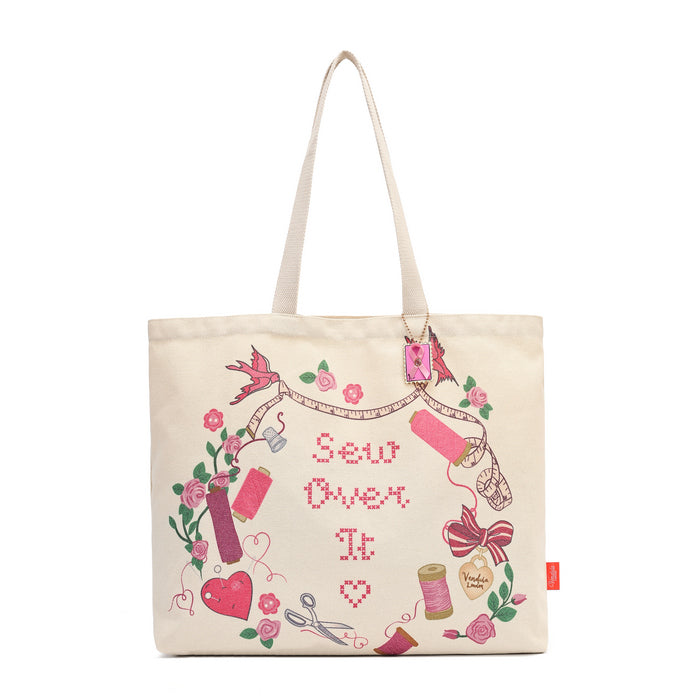 An off-white canvas tote bag with embroidered sewing-themed graphics and the text 'Sew Over It' in pink, featuring pink spools of thread, scissors, flowers, and a measuring tape.