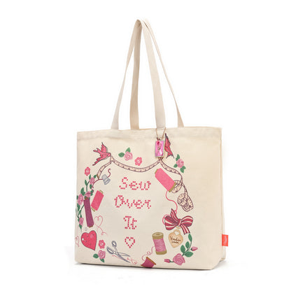 A cream canvas tote bag with long handles, decorated with a colorful 'Sew Over It' embroidery-themed graphic featuring pink, red, green, and purple details, set against a plain white background.