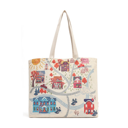 A canvas tote bag with a detailed, colorful illustrated print of a village scene, featuring houses, trees, a sun, and winding roads on an off-white background. The bag has two light beige fabric shoulder straps and is photographed against a solid white background.