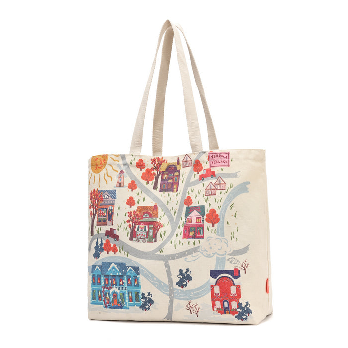 A cream-colored canvas tote bag featuring a whimsical, hand-drawn style illustration of a small village with colorful houses, trees, and winding roads.