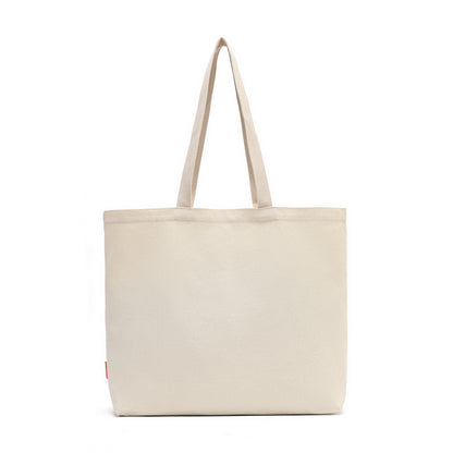 A plain canvas tote bag in a light beige or cream color, featuring two long shoulder straps and a small red tag on the lower left side, set against a solid white background.