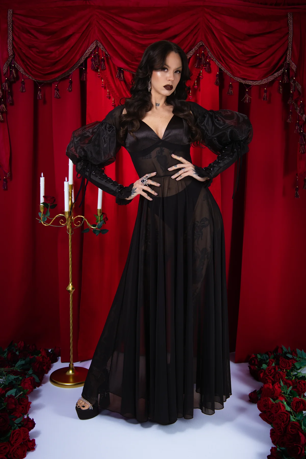 Woman in a black dress standing in front of red curtains with candles and flowers.