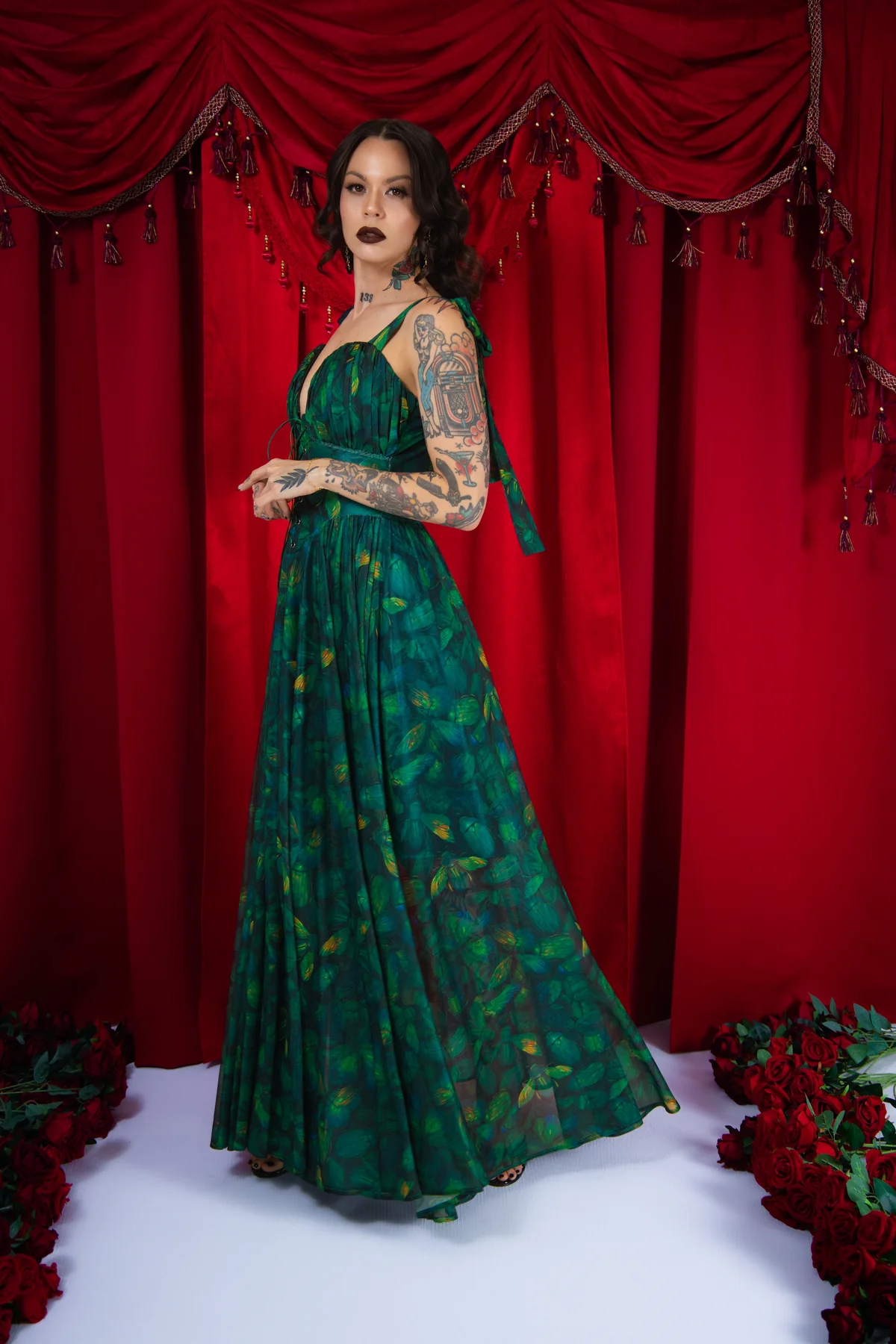 Woman in a green dress standing in front of red curtains with floral decorations on a white floor.