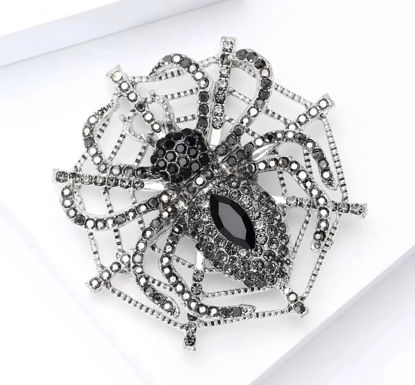 A large spider brooch featuring a black marquise-shaped center stone and black rhinestone-encrusted body, with silver-tone metal legs and web-like extensions adorned with small clear rhinestones, shown on a white background.
