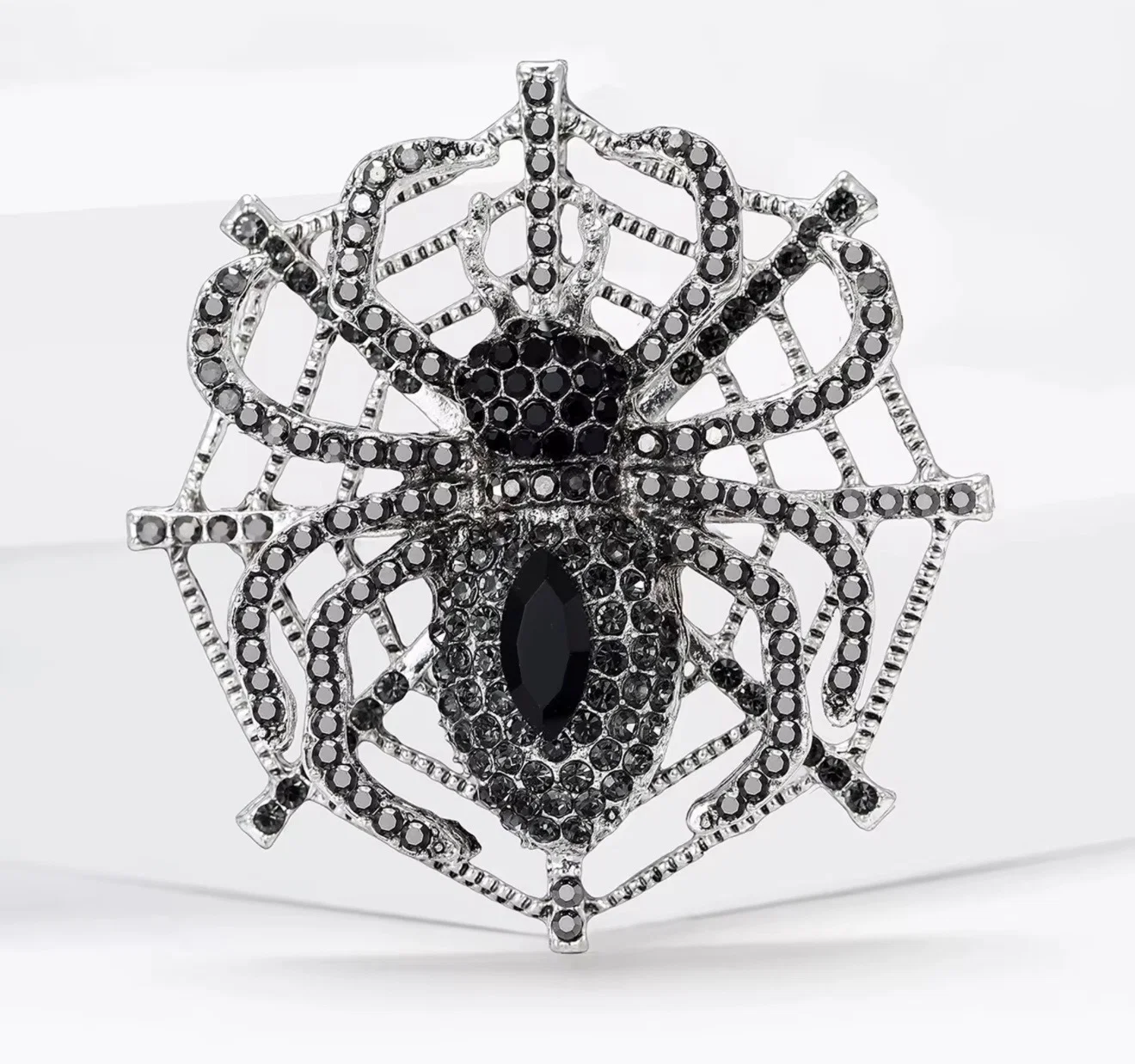 A silver-toned spider brooch embellished with black stones, featuring a large marquise-shaped black center stone and a web-like spider design, shown on a white background.