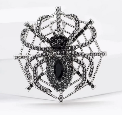 A silver-toned spider brooch embellished with black stones, featuring a large marquise-shaped black center stone and a web-like spider design, shown on a white background.