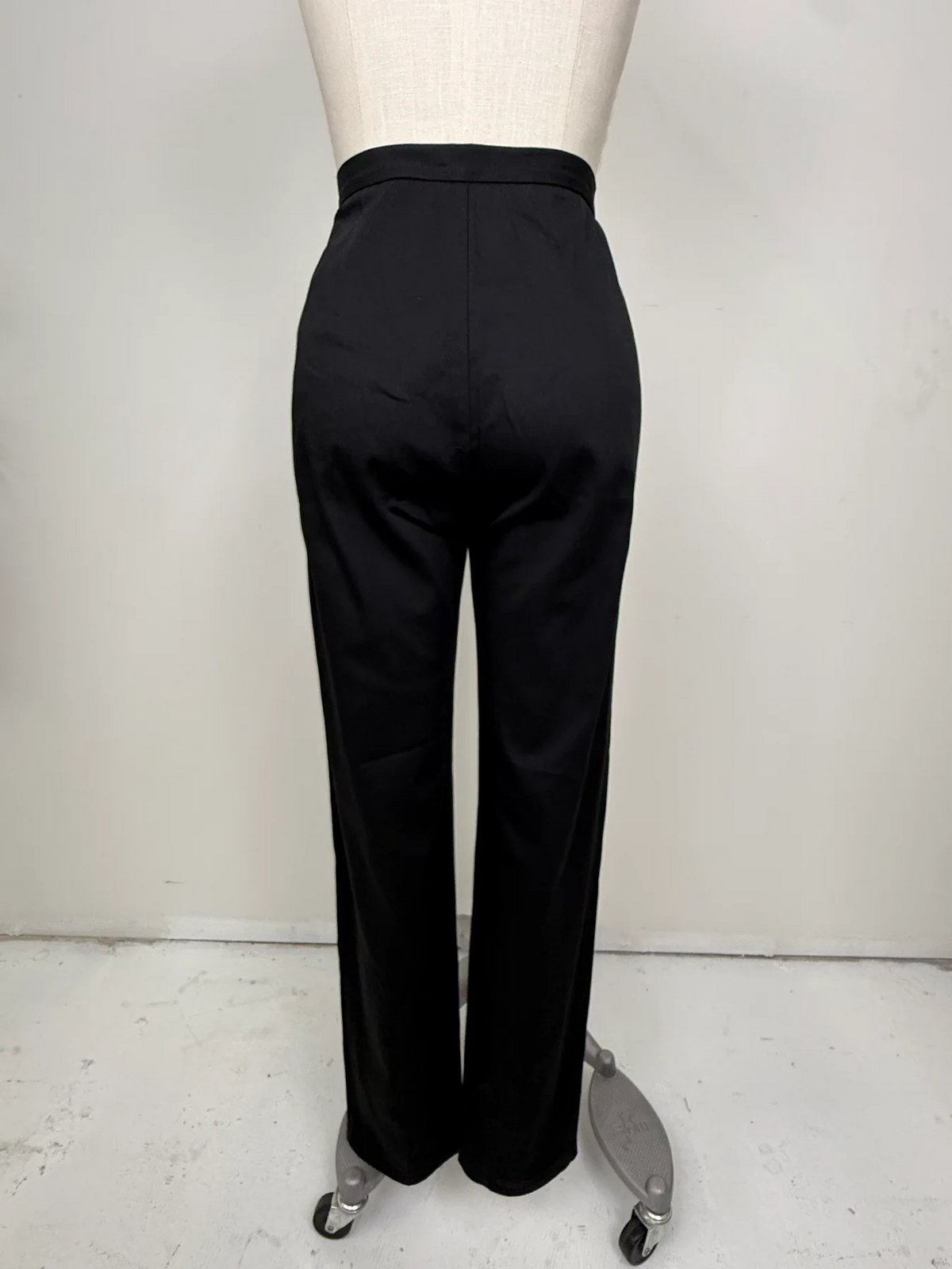 Black wide-leg trousers shown from the back on a dress form mannequin against a plain light gray wall.