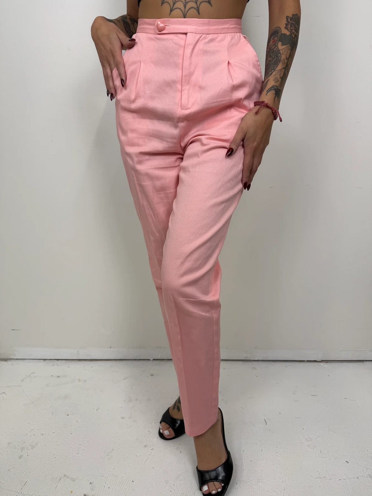 A person modeling a pair of light pink high-waisted cropped trousers with front pleats and side pockets, paired with black heeled sandals against a plain white wall.