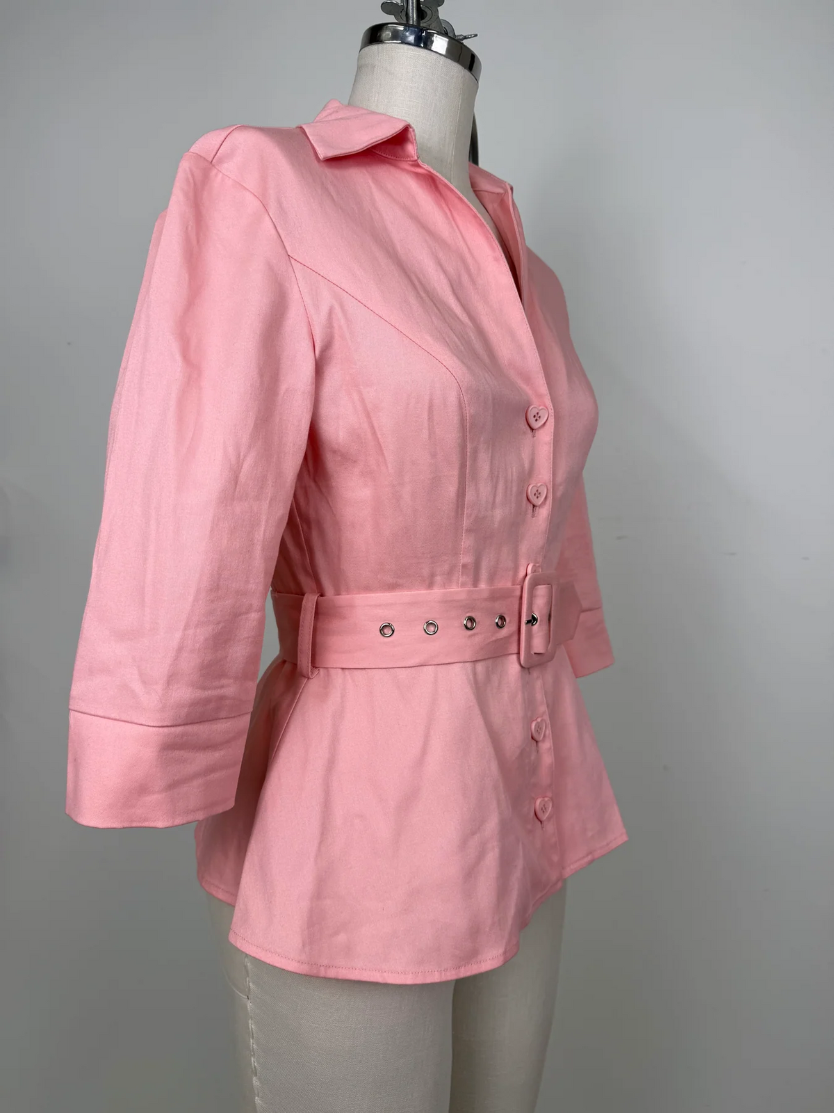 A pink women's blazer-style jacket with a collar, three-quarter sleeves, front buttons, and a matching waist belt with metal eyelets, displayed on a dress form against a plain light gray background.