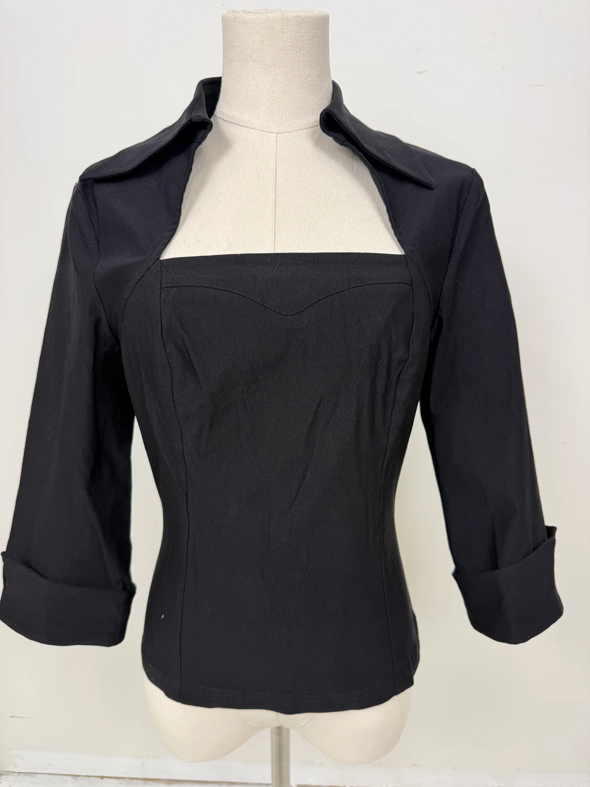 A black long-sleeve blouse with a square neckline and structured front panel, displayed on a cream mannequin torso against a plain white background.