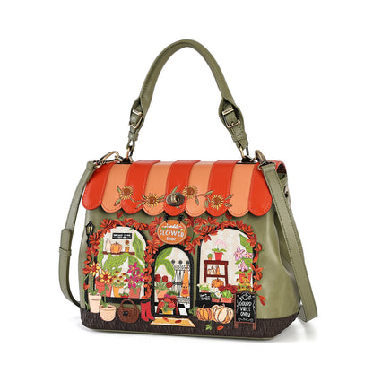 An olive green handbag with a detailed autumn-themed floral storefront patchwork design on the front, featuring a scalloped orange and peach roof, floral arches, pumpkins, and a hanging shoulder strap.